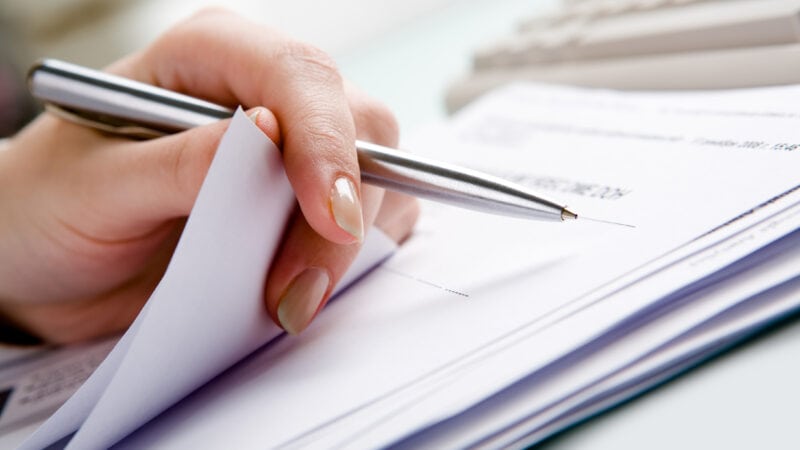 Close-up of  hand holding pen with paper over pile of documents