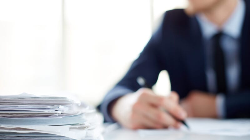 Stack of documents at workplace and male employee on background